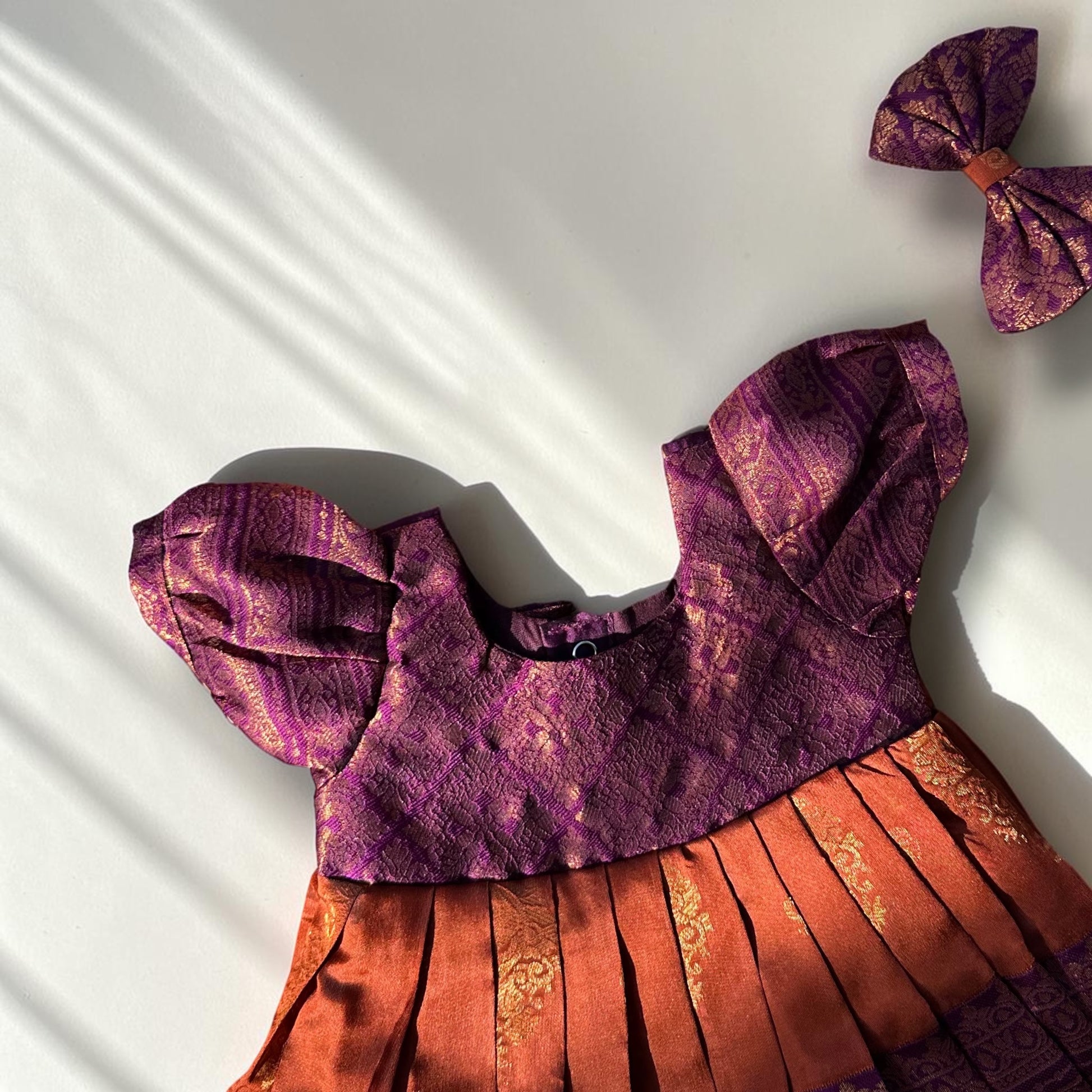 A purple and orange patterned traditional dress laid flat on a white background with diagonal shadows, accompanied by a matching bow accessory in the upper right corner.