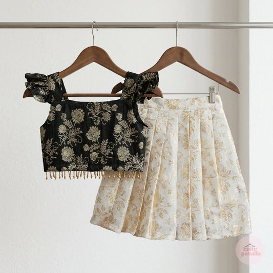 A two-piece girls' outfit hanging on wooden hangers against a white wall. The set includes a black floral off-the-shoulder crop top with gold fringe and a matching pleated skirt in cream with a gold floral pattern.