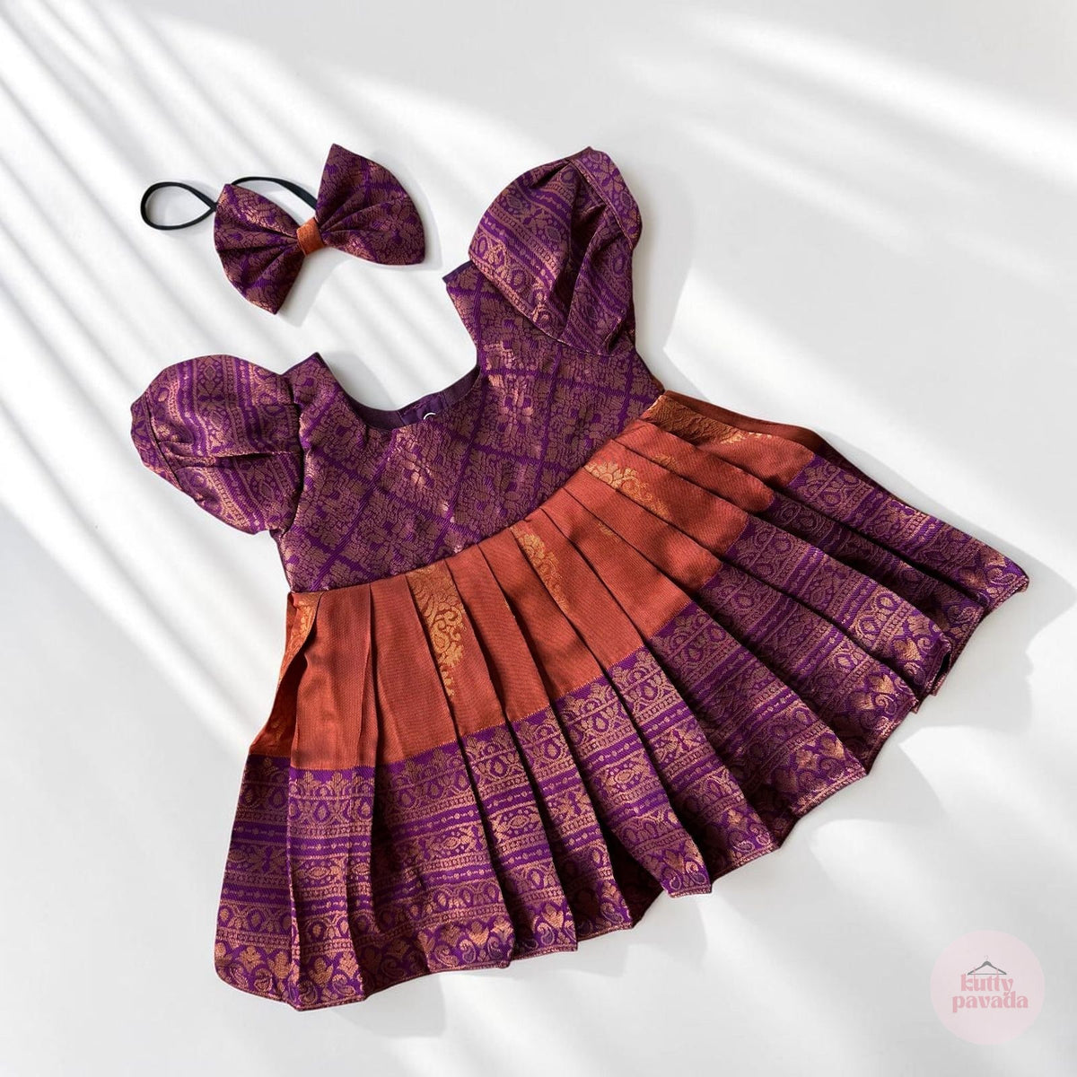 A baby girl's traditional Indian outfit set featuring a purple and rust-orange patterned dress with a pleated skirt, accompanied by a matching bow headband, laid flat on a white background with soft leaf shadows.