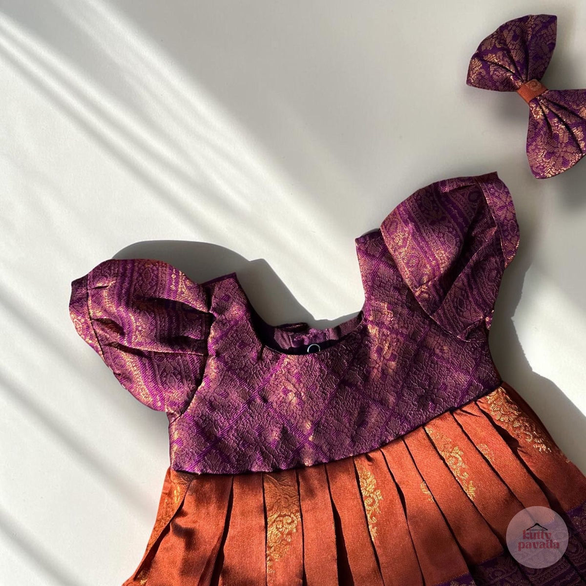 A purple and orange patterned traditional dress laid flat on a white background with diagonal shadows, accompanied by a matching bow accessory in the upper right corner.