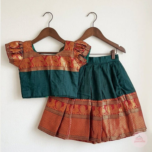 A two-piece traditional outfit consisting of a short-sleeve blouse and a pleated skirt, hanging on wooden hangers against a plain white wall. The outfit features a dark teal green upper section and a rust-orange lower section with gold zari embroidery.