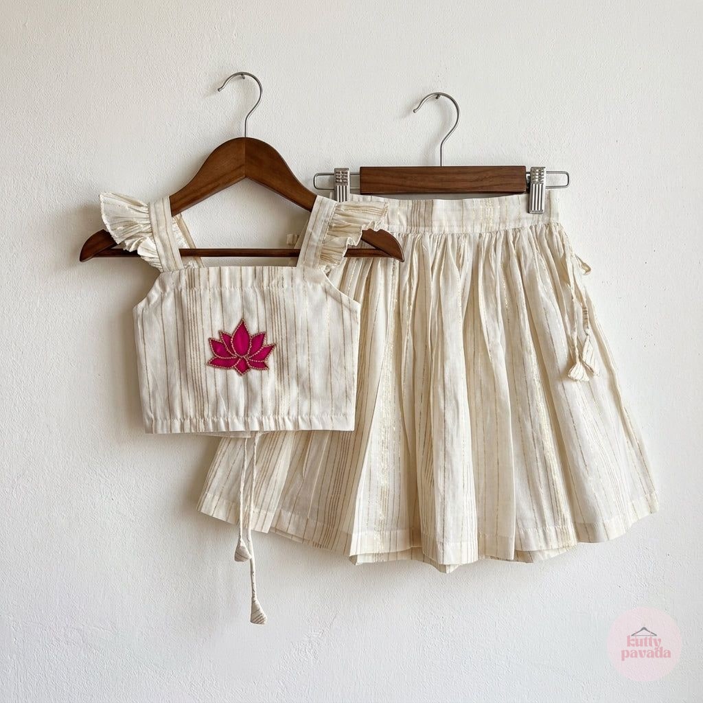 A two-piece children's outfit set consisting of a cream-colored linen-look crop top with ruffle straps and a matching pleated skirt, hanging on wooden hangers against a white wall. The crop top features a pink embroidered maple leaf on the chest.