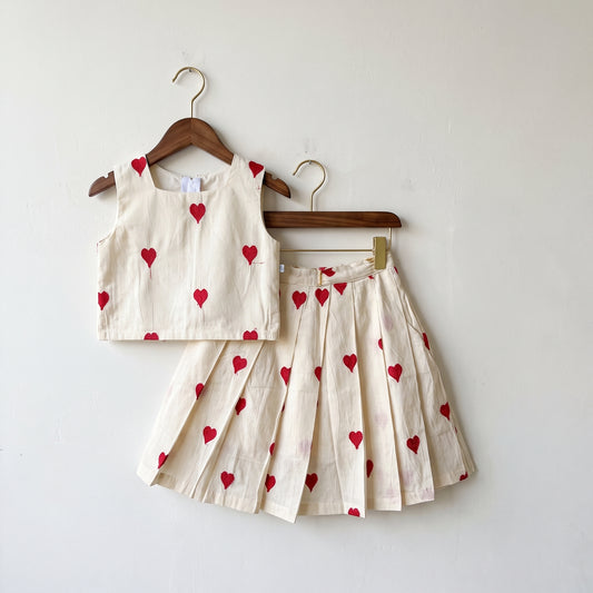 A two-piece children's outfit set consisting of a sleeveless crop top and a pleated skirt, both in a light beige linen fabric with a red heart print, hanging on wooden hangers against a plain white wall.