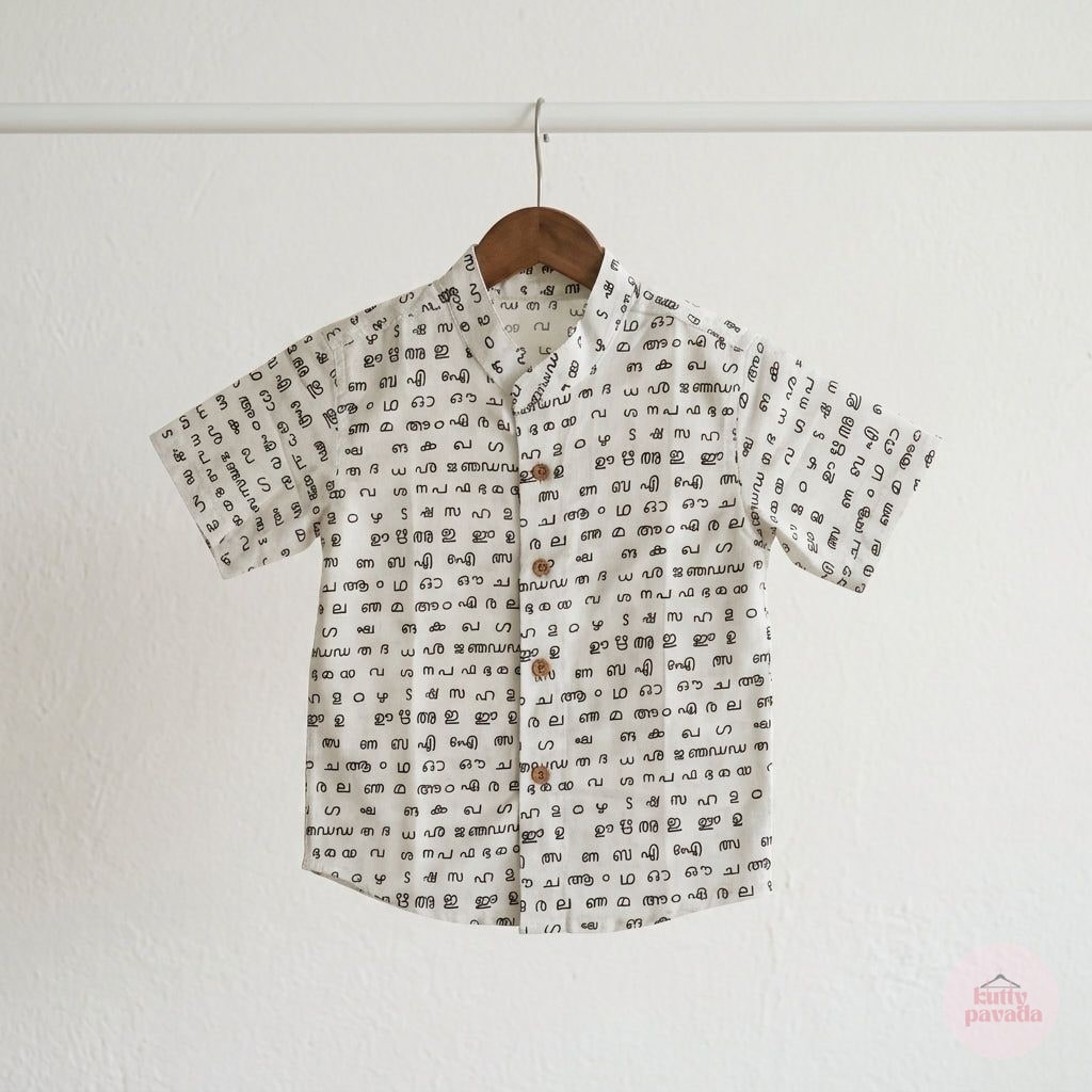 A short-sleeve button-up shirt with an all-over black text pattern on a white background, hanging on a wooden hanger against a white wall.