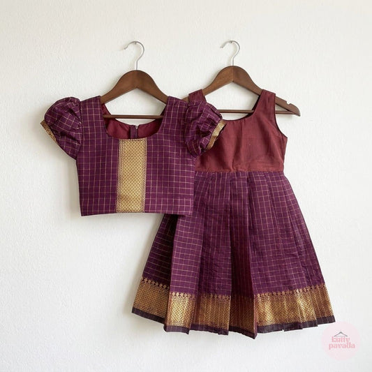 A two-piece toddler girl's traditional outfit hanging on wooden hangers against a white wall. The set includes a sleeveless top with puff sleeves and a matching pleated skirt, both in a deep purple and gold checkered fabric with a solid maroon bodice.