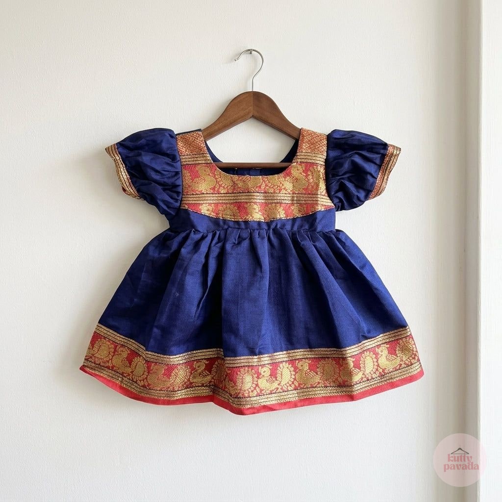 A navy blue baby dress with gold and red traditional patterns, hanging on a wooden hanger against a plain white wall.