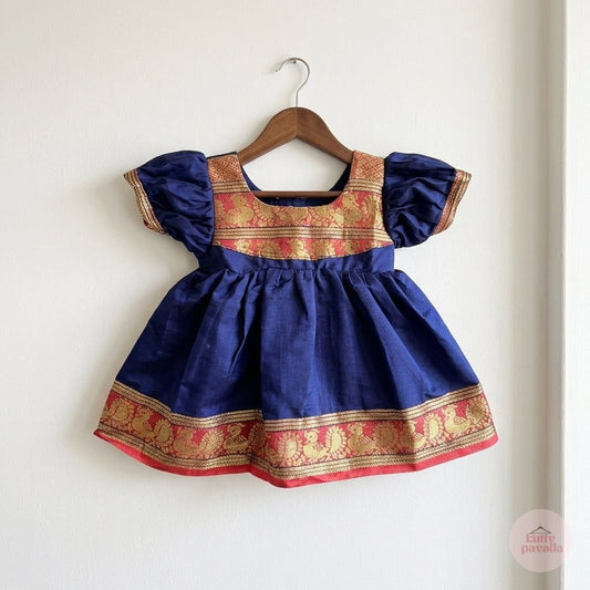 A navy blue baby dress with gold and red traditional patterns, hanging on a wooden hanger against a plain white wall.