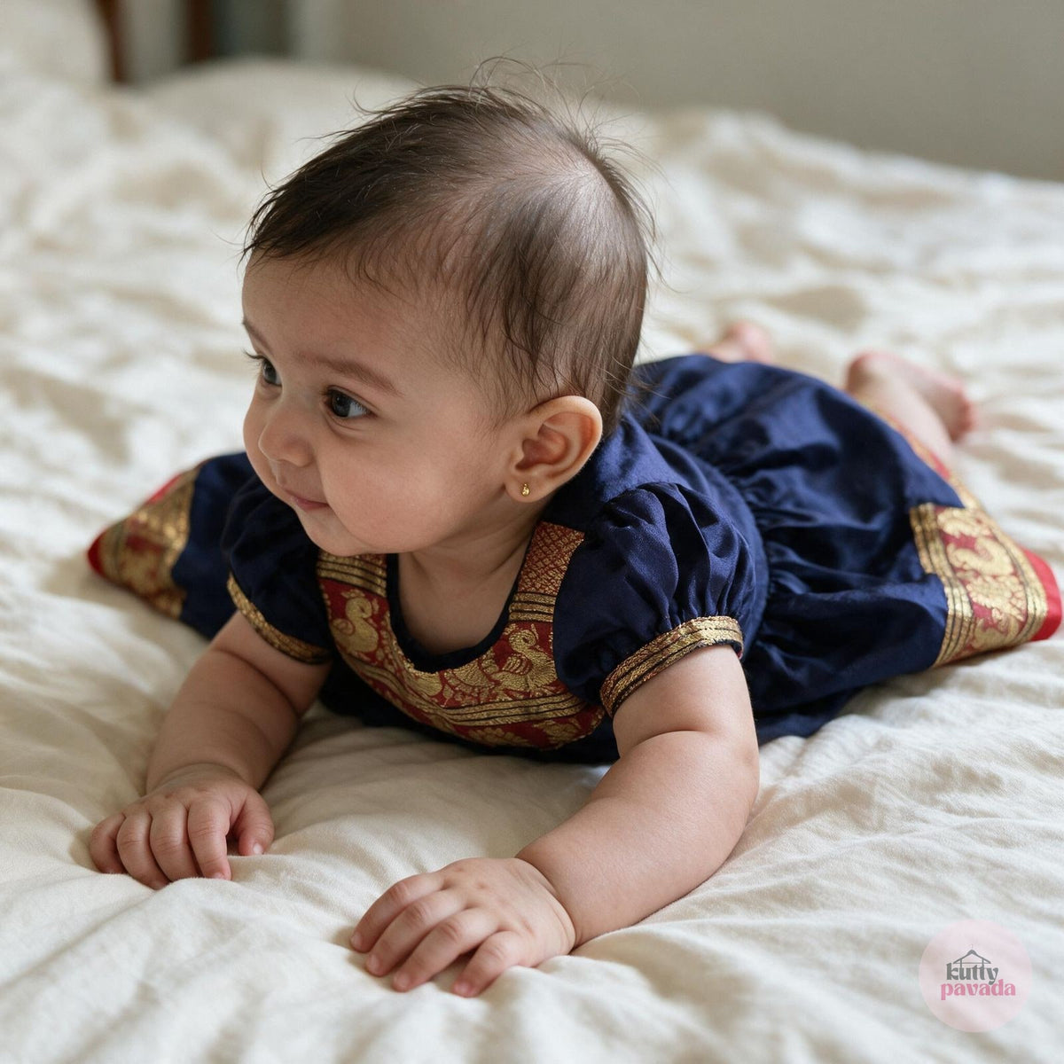 A baby girl during tummy time on a cream bedsheet, wearing a navy blue silk frock with a red and gold brocade yoke and matching hem border.