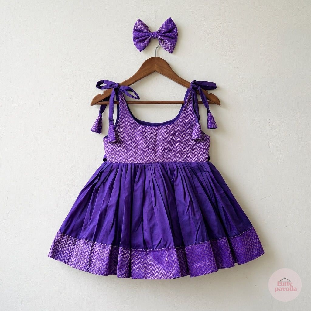 A purple children's dress with a matching bow headband, displayed on a wooden hanger against a plain off-white wall. The dress features a chevron-patterned bodice and upper skirt, a solid purple gathered waist, and a ruffled hem with a matching chevron pattern.