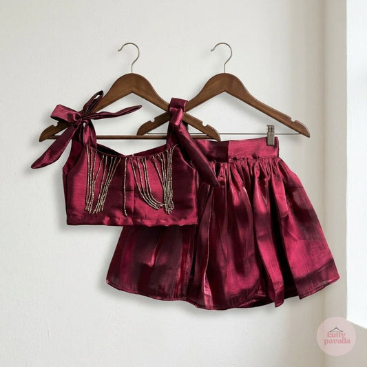 A two-piece girls' outfit set in a shiny deep burgundy satin fabric, hanging on wooden hangers against a plain off-white wall. The set includes a tie-shoulder crop top with dangling beaded fringe and a matching pleated mini skirt.