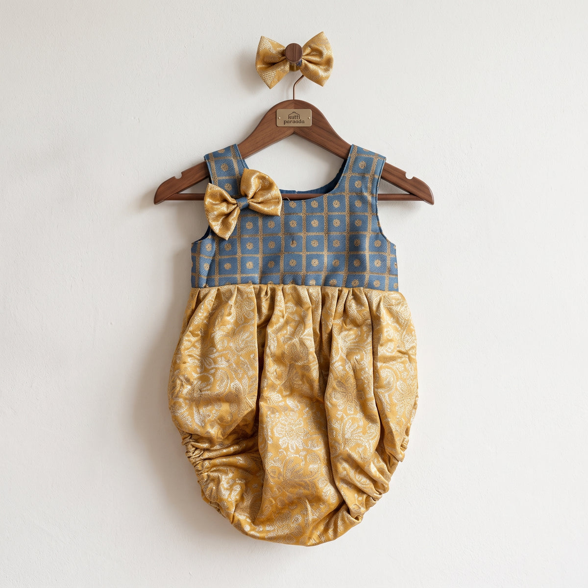 A baby girl's sleeveless romper dress hanging on a wooden hanger against a plain white wall. The romper features a dusty blue patterned bodice with a matching gold bow on the shoulder, and a gathered gold satin bubble bottom with a subtle damask pattern.