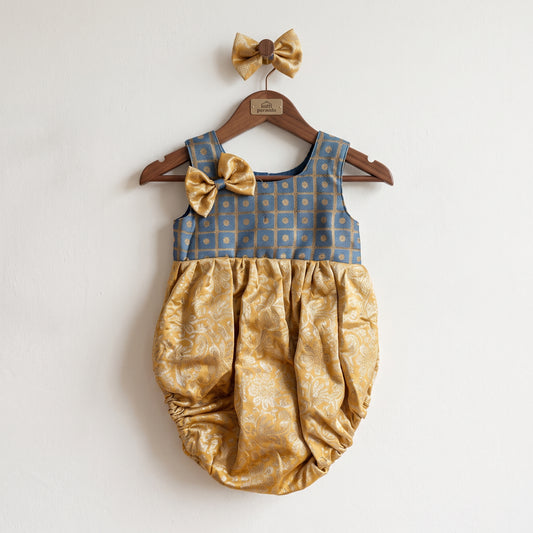 A baby girl's sleeveless romper dress hanging on a wooden hanger against a plain white wall. The romper features a dusty blue patterned bodice with a matching gold bow on the shoulder, and a gathered gold satin bubble bottom with a subtle damask pattern.