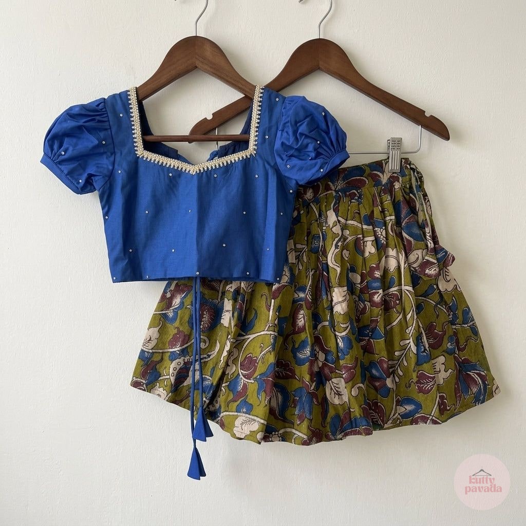 A two-piece children's outfit hanging on wooden hangers against a plain off-white wall. The set includes a royal blue short-sleeve crop top with puff sleeves and a square neckline trimmed with cream embroidery, paired with a pleated olive green skirt featuring a multicolor floral and vine print.