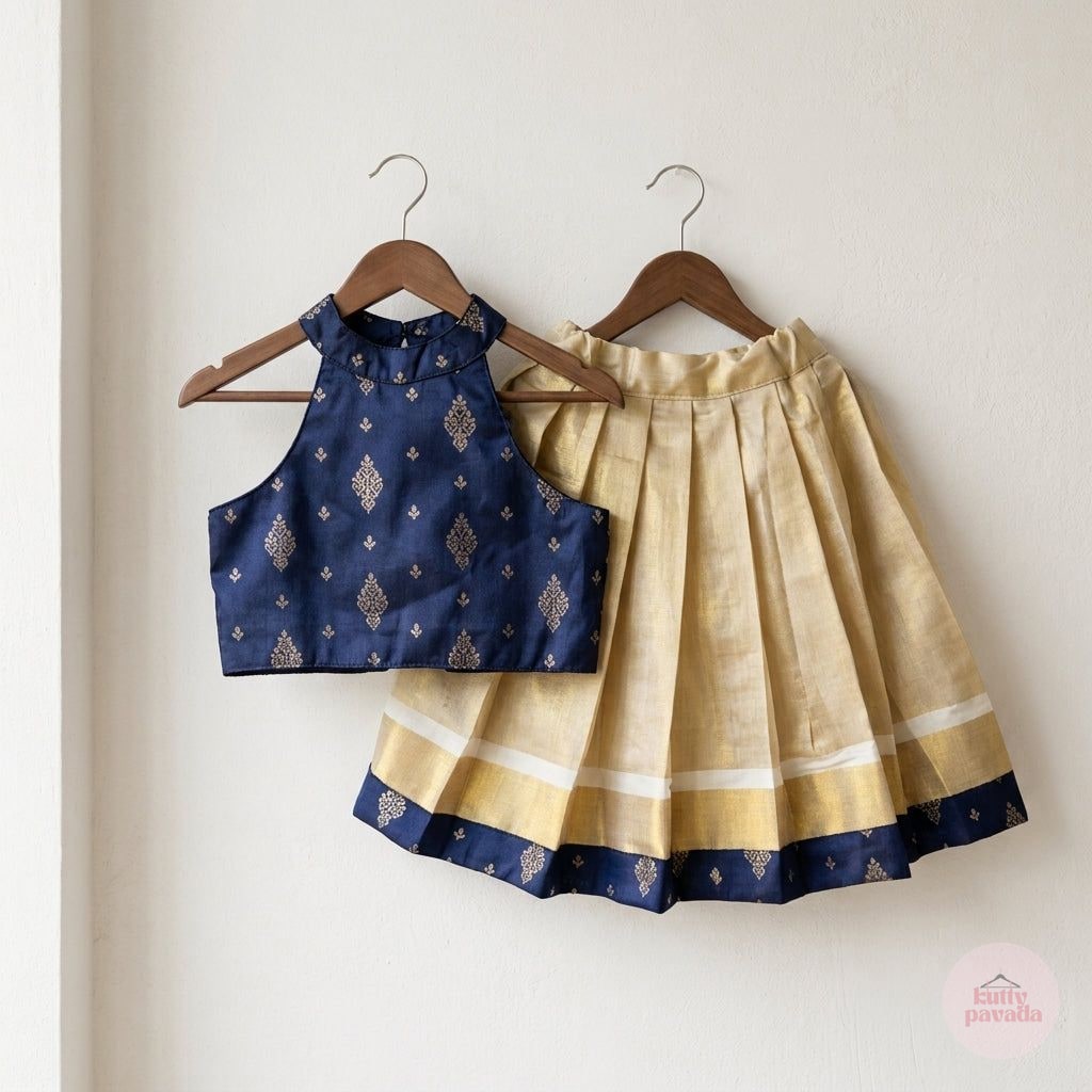 A two-piece children's traditional outfit hanging on wooden hangers against a plain off-white wall. The set includes a navy blue sleeveless top with gold motifs and a pleated skirt in beige and navy with white stripes and a navy border.