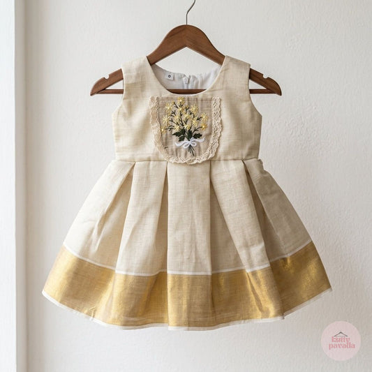 A sleeveless baby girl's dress in natural linen fabric, featuring a gold metallic border along the hem and a decorative embroidered floral chest panel, hanging on a wooden hanger against a white wall.