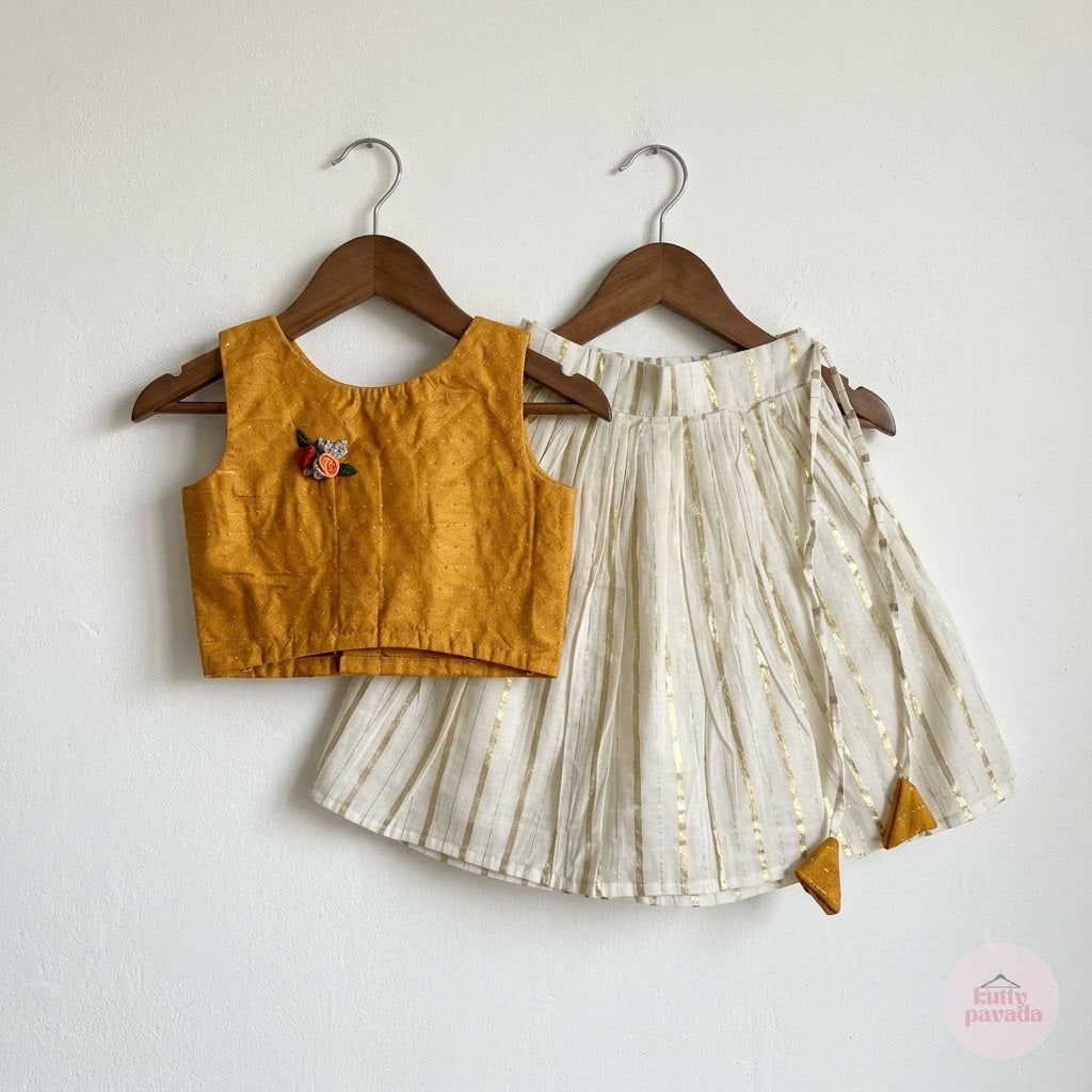 A two-piece children's outfit hanging on wooden hangers against a white wall. The set includes a mustard yellow sleeveless crop top with a small floral embroidery on the chest, and a pleated off-white skirt with thin gold vertical stripes and small gold corner accents.
