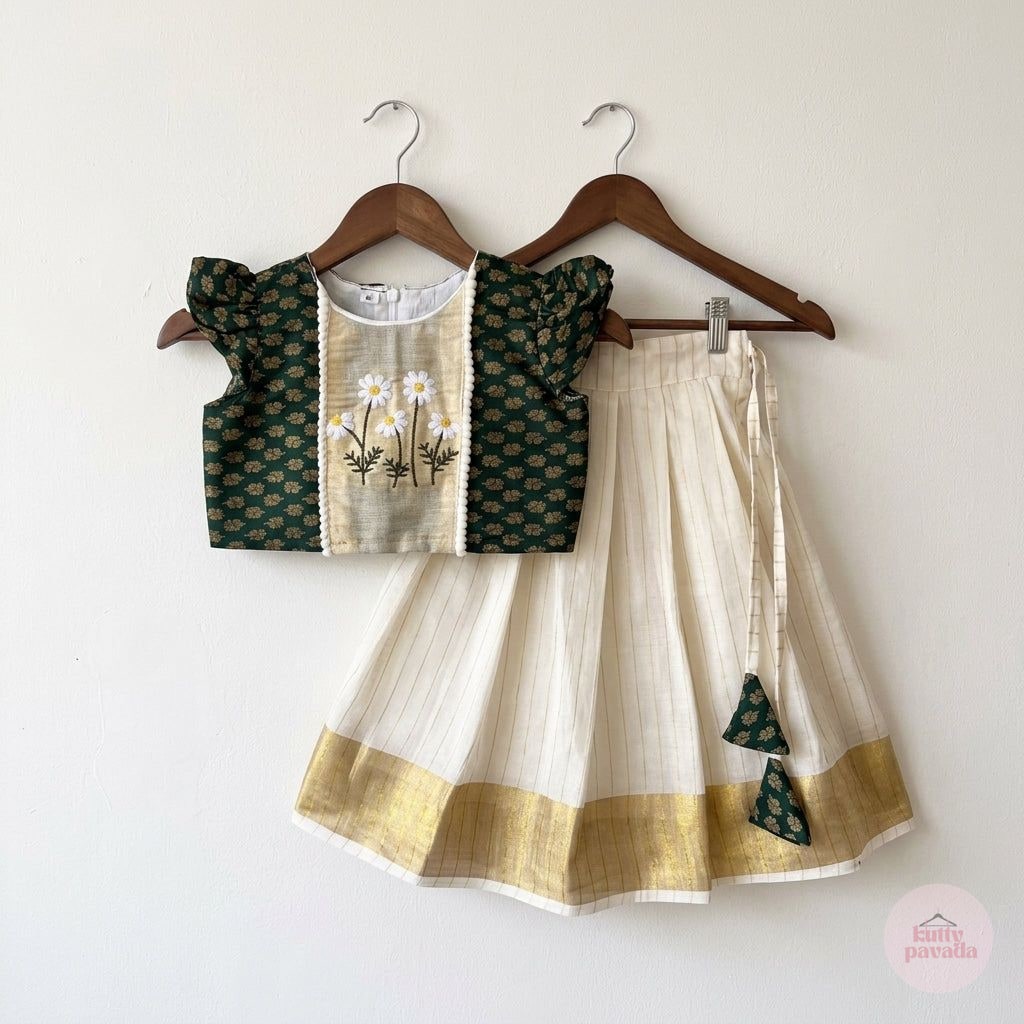 A two-piece children's traditional outfit hanging on wooden hangers against a plain off-white wall. The set includes a dark green patterned sleeveless top with a beige front panel featuring embroidered daisies, and a cream pleated skirt with a gold border and green patterned tie details.