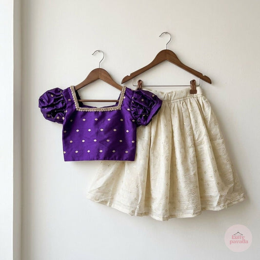 A two-piece children's outfit hanging on wooden hangers against a plain off-white wall. The set includes a purple short-sleeve blouse with gold polka dots and a square neckline, paired with a cream-colored textured flared skirt.