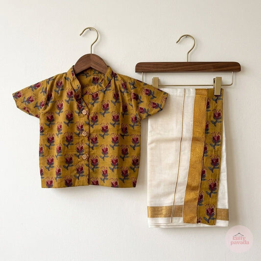 A two-piece children's outfit hanging on wooden hangers against a plain white wall. The set includes a mustard yellow short-sleeve button-up shirt with a red and green floral block print, and a white dhoti-style bottom with a wide gold stripe and matching floral print.